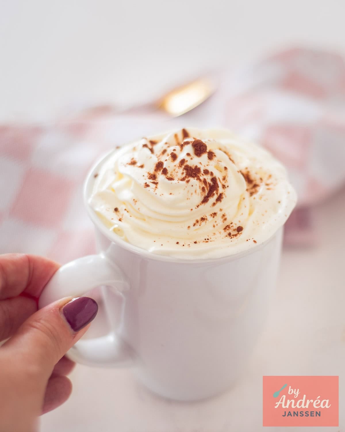 A hand holding a cup of white hot chocolate with whipped cream and cocoa on top.