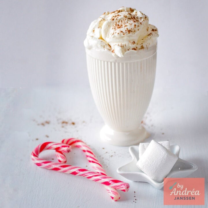 A square picture with white hot chocolate topped with whipped cream. Some marshmallows in front of the mug.