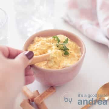 A square photo of a pink container filled with an egg salad without mayonnaise. A tapas stick scoops something out. A white background. Top right a pink white tea towel
