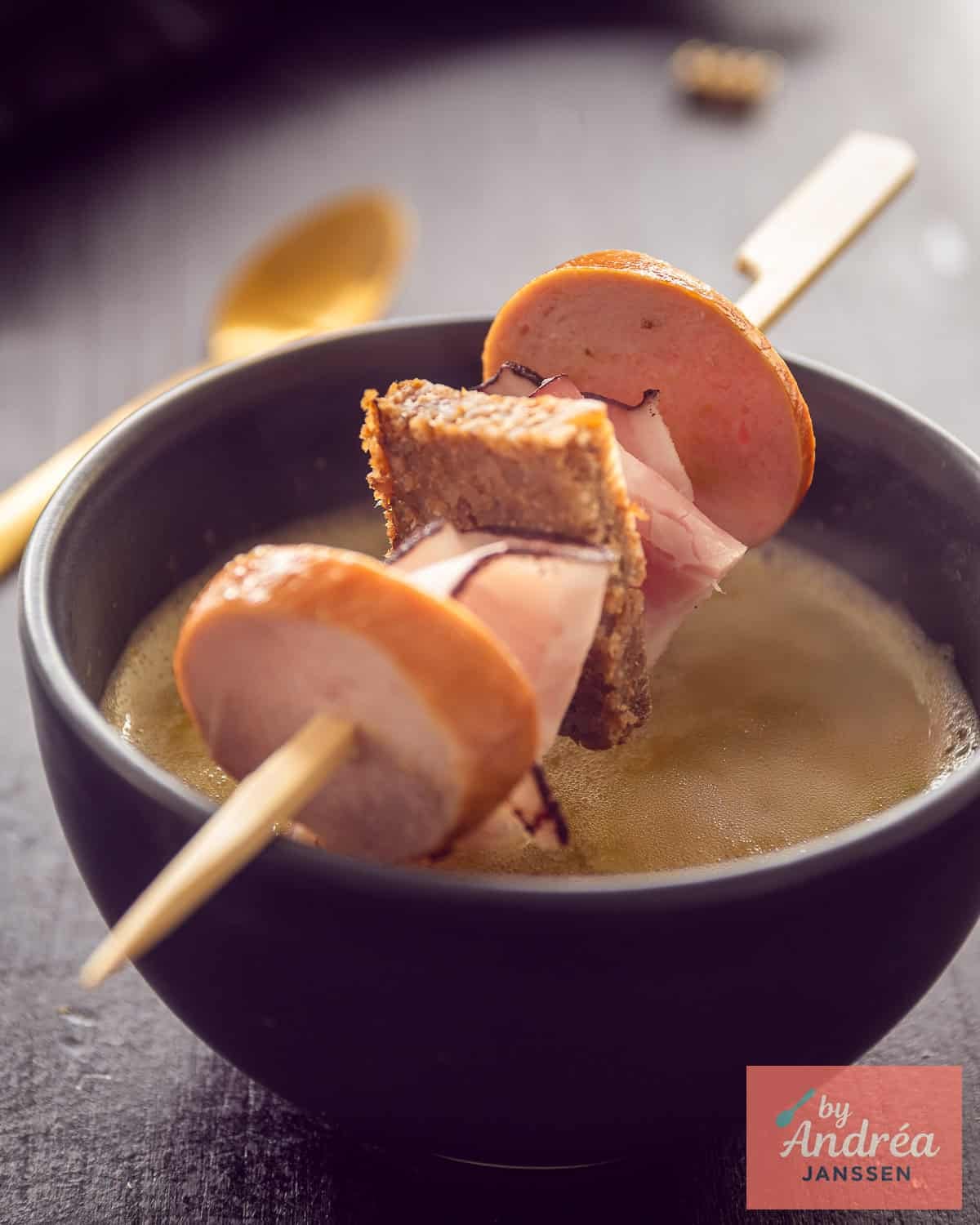 Een zwarte kom met snert, en een stokje met rookworst, spek en roggebrood erboven.