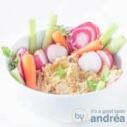 A square photo with a white bowl filled with hummus with carrots, radishes and celery in it.
