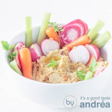 A square photo with a white bowl filled with hummus with carrots, radishes and celery in it.