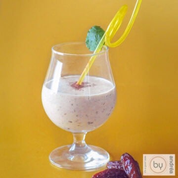 A square photo with a yellow background. In the foreground a mango coconut smoothie with dates.