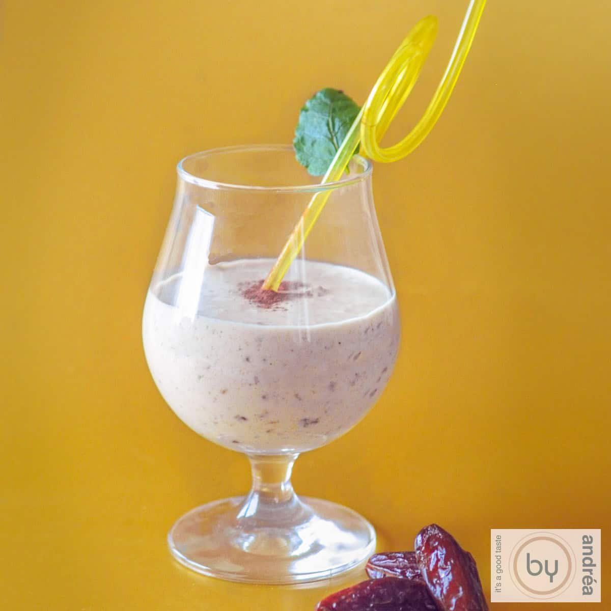 A square photo with a yellow background. In the foreground a mango coconut smoothie with dates.