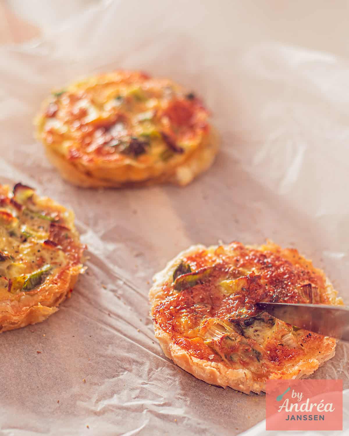 Three mini quiches on a baking sheet. A knife cuts into one of the puff pastry quiches with smoked mackerel, leek, and cheese.