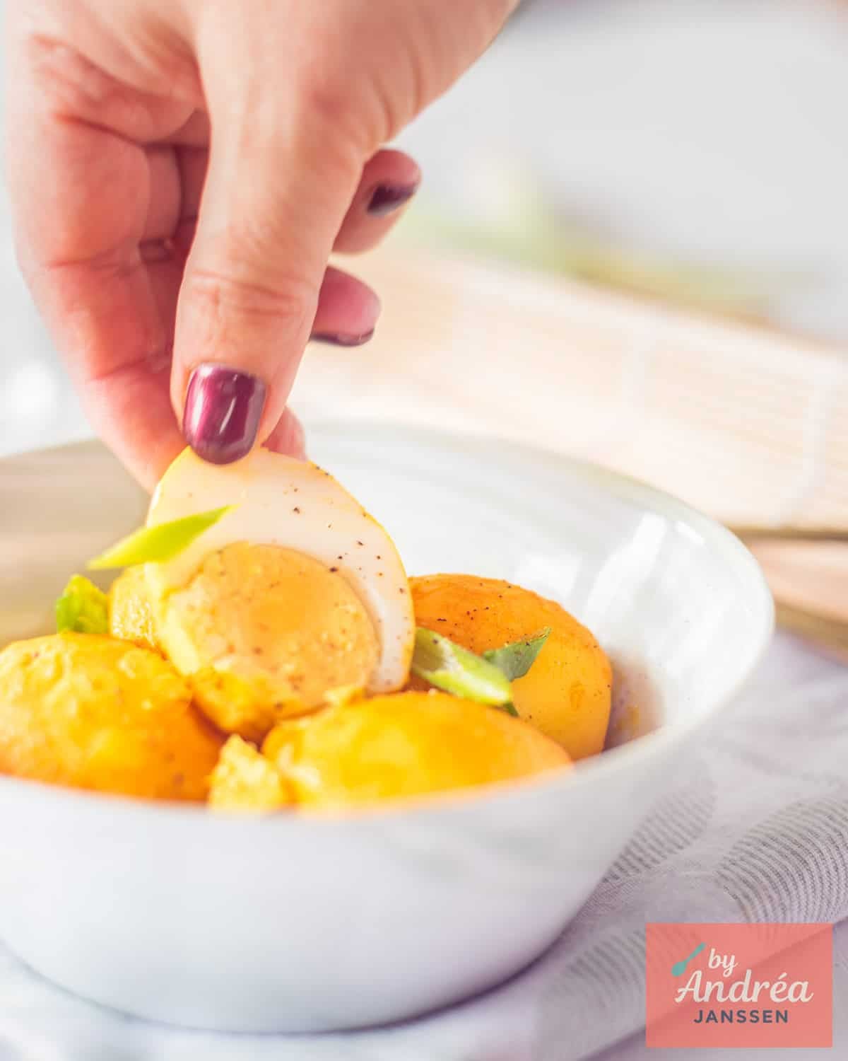 A hand takes an egg stewed in coconut milk with galangal and lemongrass from a green bowl.