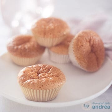 A square photo with a white plate with five sweet milk cupcakes, one in the foreground. A white background.