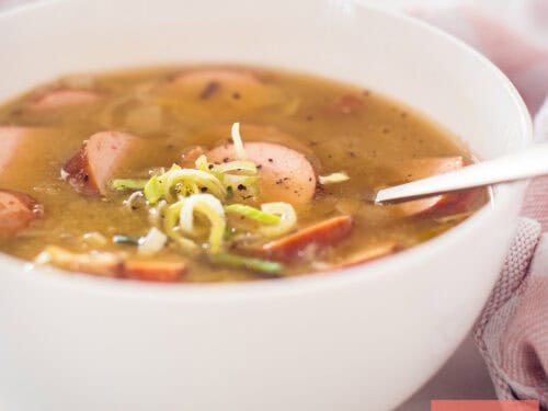 A square photo of a white bowl of pea soup with sausage and ham, just like grandma makes.