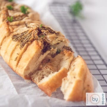 A square photo with a baguette on a baking sheet, with two slices cut off and the garlic oil and herbs visible.