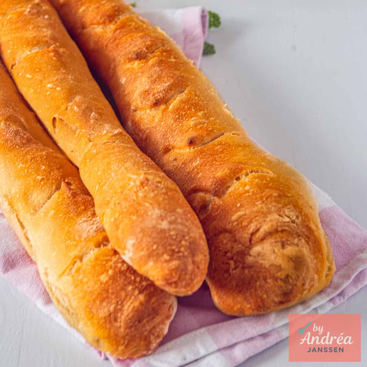 A square photo with three homemade baguettes in a stack on a tea towel.