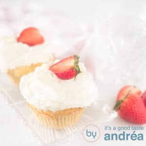 A square photo of two cupcakes on a white surface on a bowl with a dollop of whipped cream and a strawberry on top