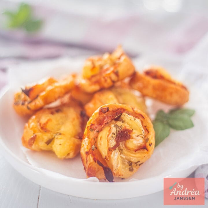 A square photo with a white plate full of puff pastry bites with cheese and bacon.