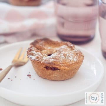 A square photo with a white plate with a mini apple pie on it. In the background pink glassware and a pink white tea towel