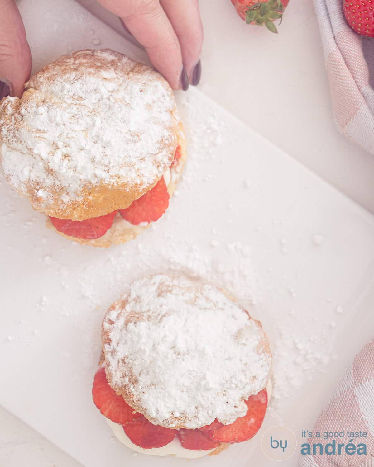 Een foto van bovenaf van twee scones met aardbeien en room. Een hand pakt de bovenste scone.