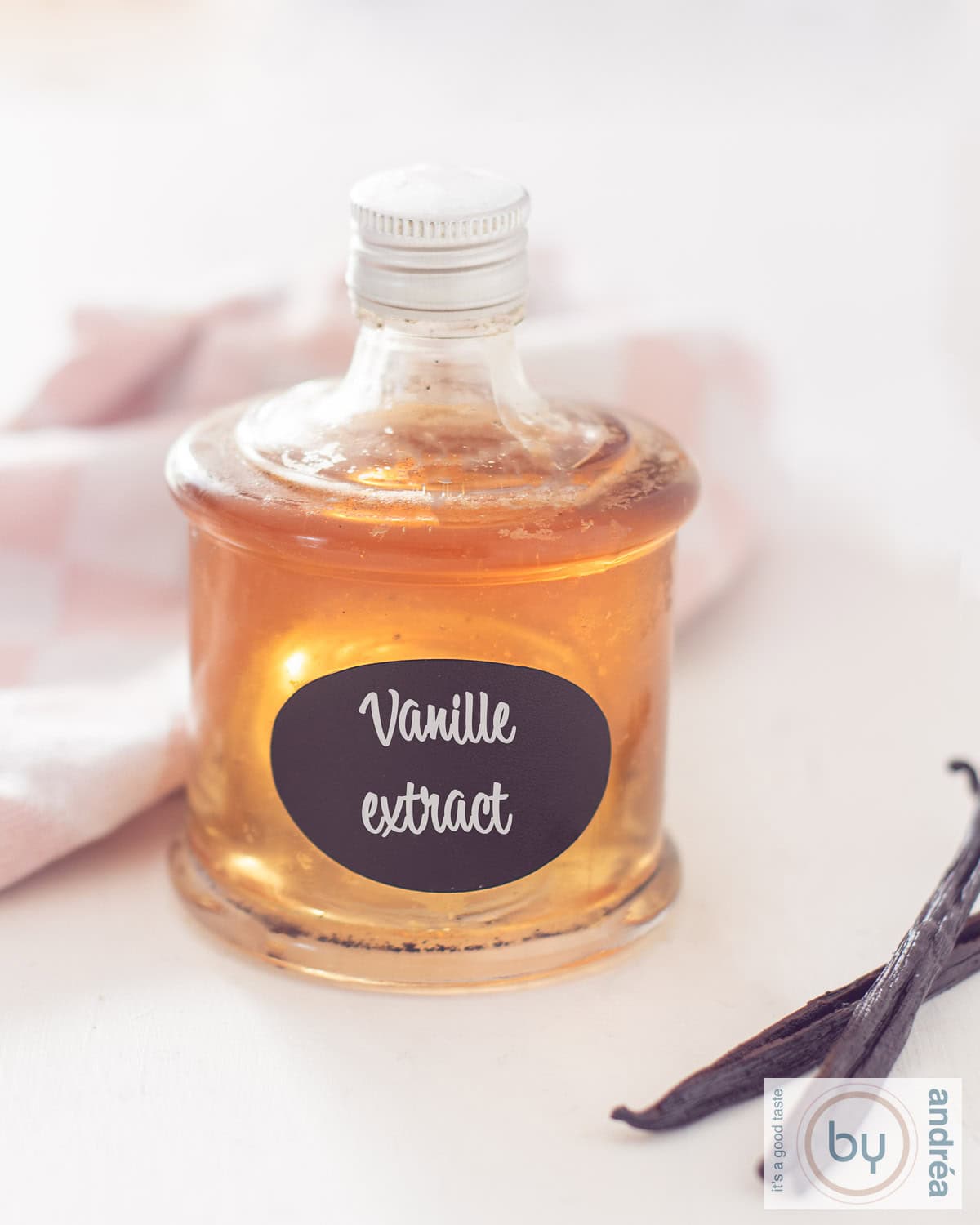 Een hoogtefoto met een fles gevuld met vanille extract op een witte ondergrond. Rechtsachter een roze witte theedoek, linksvoor twee vanillestokjes