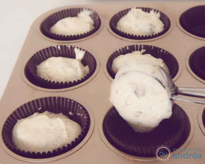 A baking tray with brown paper cases that are filled with cupcake batter with an ice cream scoop.