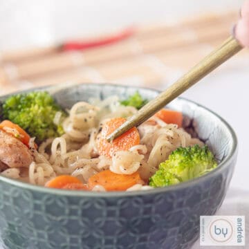 A square picture with a green bowl filled with noodles, carrots broccoli and chicken. Chopsticks taking a carrot.