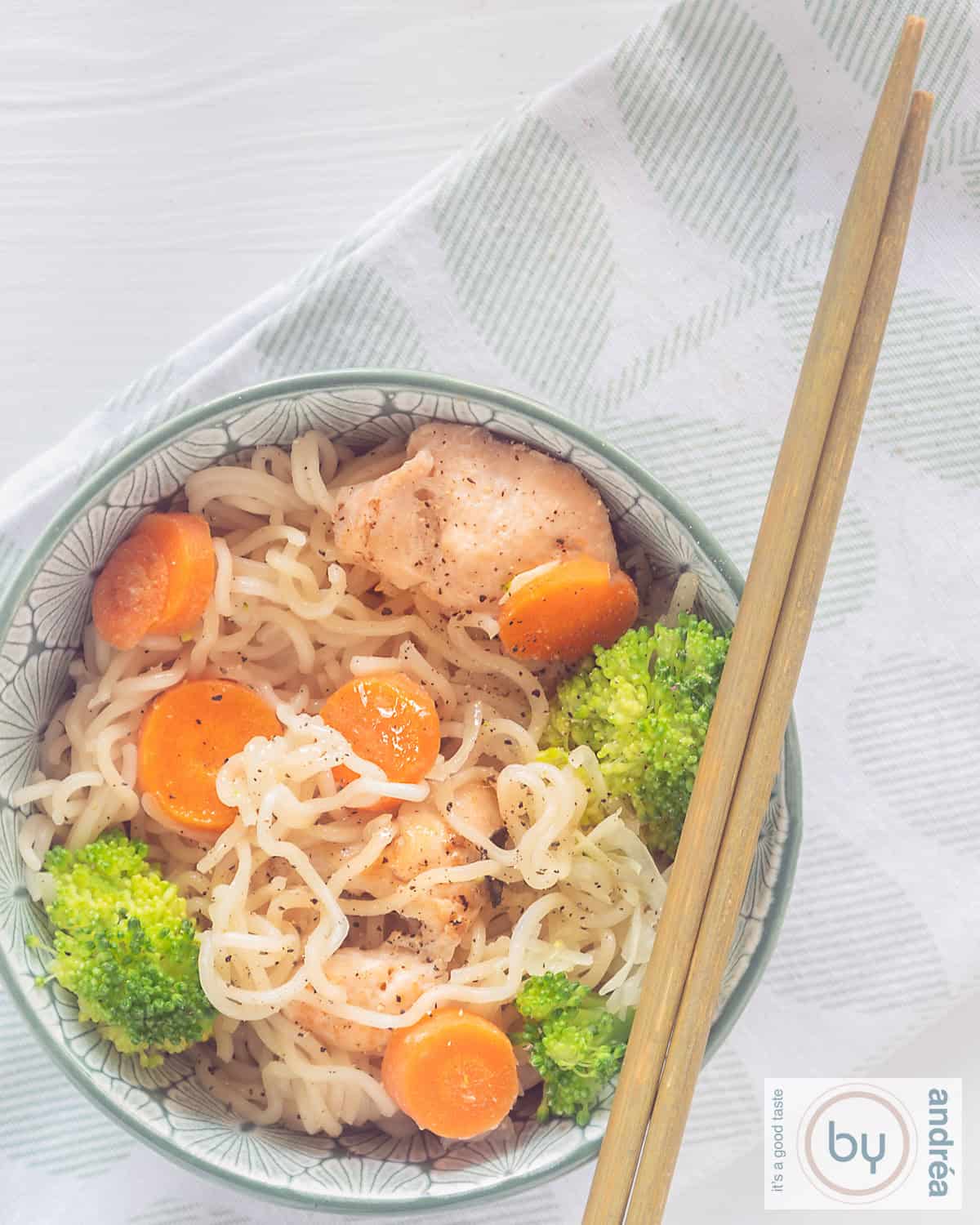 A top down picture a bowl filled with noodles, chicken, carrots and broccoli. Two chopsticks laying on the bowl
