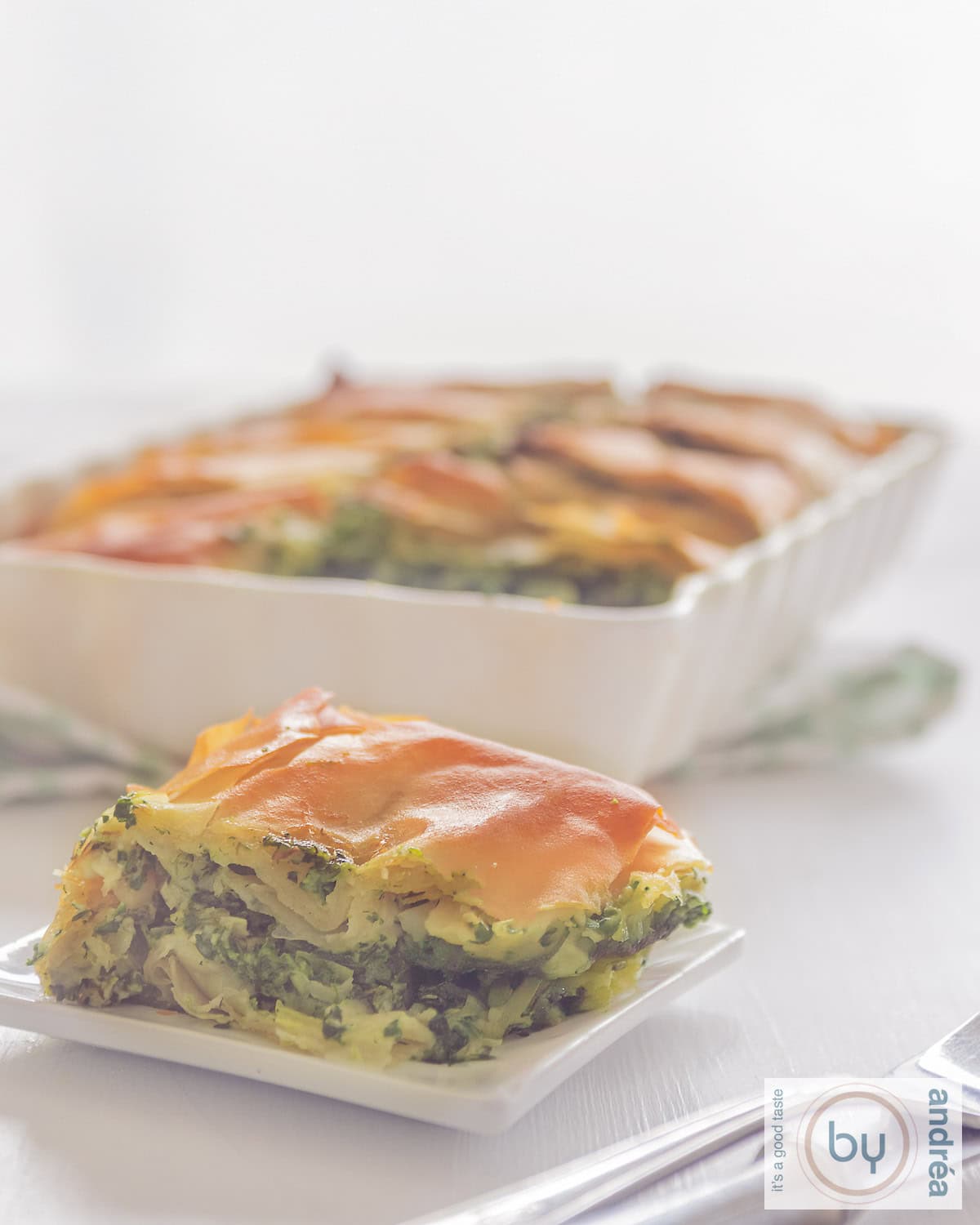 Een foot met een bord met een stuk spanakopita. Een ovenschaal met meerdere stukjes in de achtergrond.