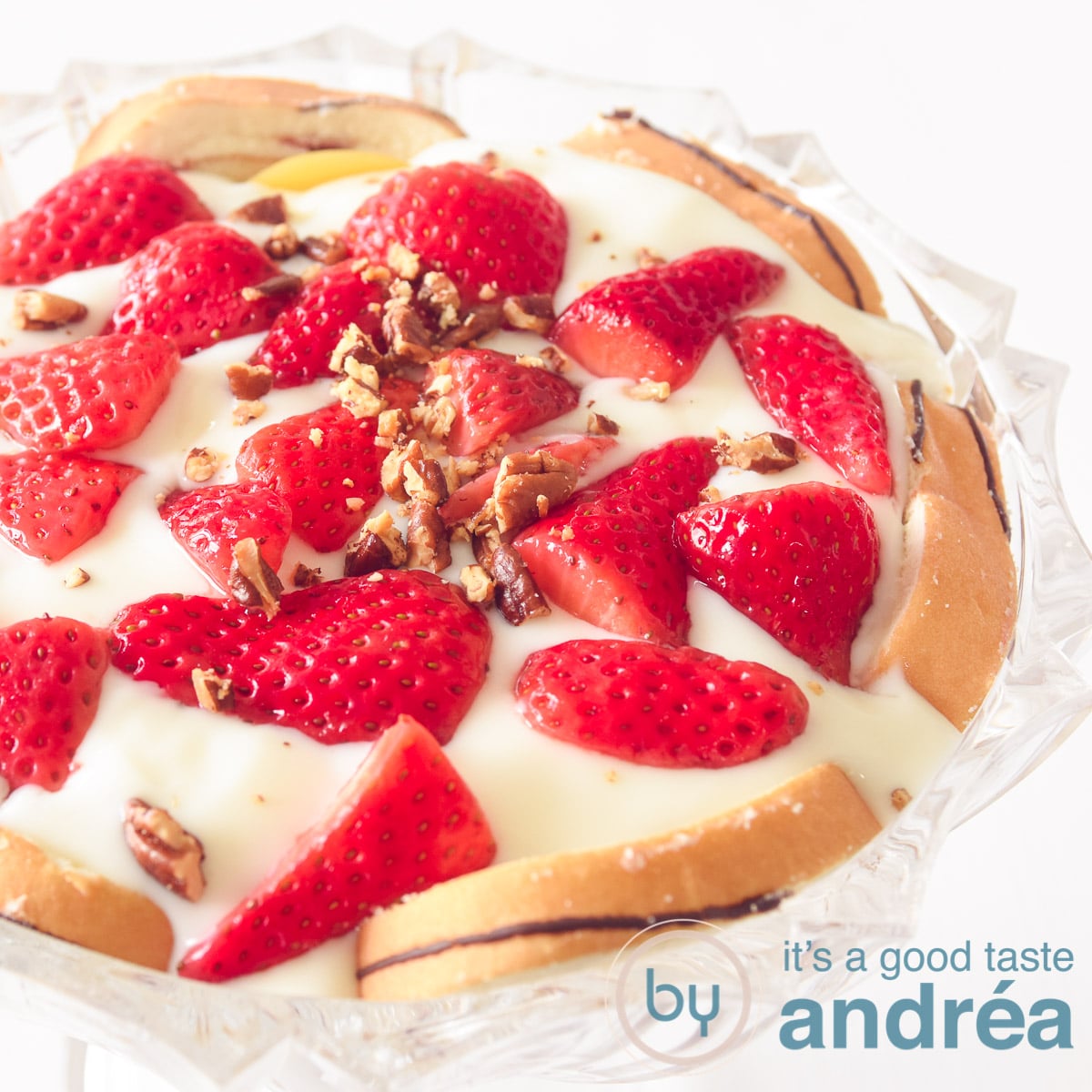 A square photo with a glass bowl filled with layers of cake, cream, strawberries and pecans