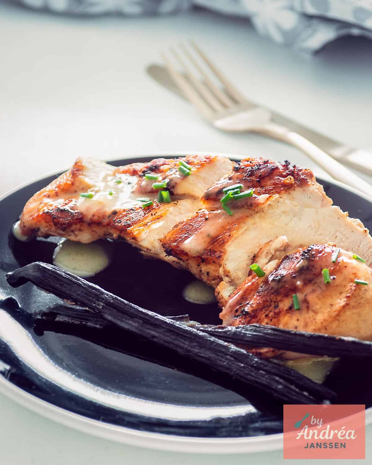 A black plate with sliced ​​fried chicken breast, vanilla sauce, and a vanilla pod.