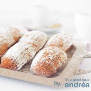A square photo with an aluminum bowl with madeleines with powdered sugar on a white background