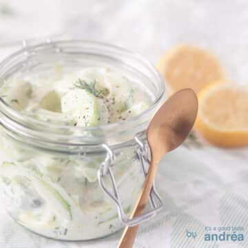A square photo with a glass jar containing a cucumber salad with Greek yogurt. There's a spoon in there for it. Two lemons in the top right.