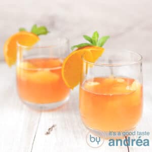 A square photo with two glasses with orange elderflower cocktail on a wooden floor.