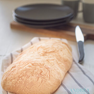 A loaf of homemade ciabatta on a towel with a knife, plates and mugs