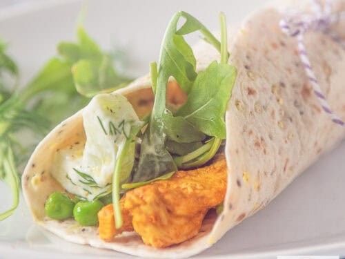 A square photo of a white plate with a rolled wrap filled with Tandoori chicken, tzatziki and vegetables