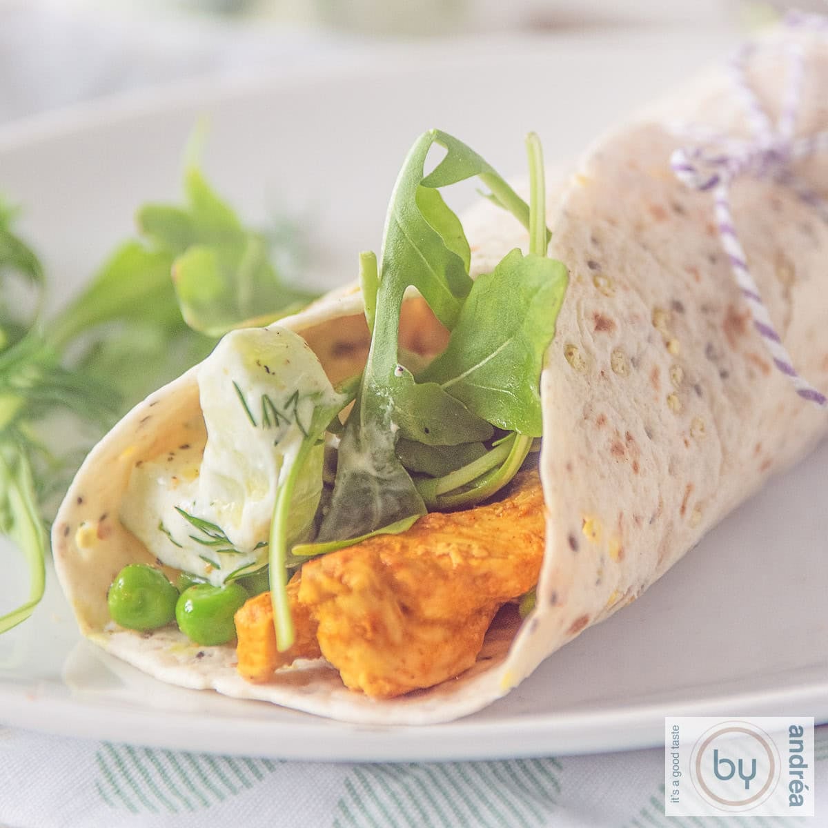 A square photo of a white plate with a rolled wrap filled with Tandoori chicken, tzatziki and vegetables