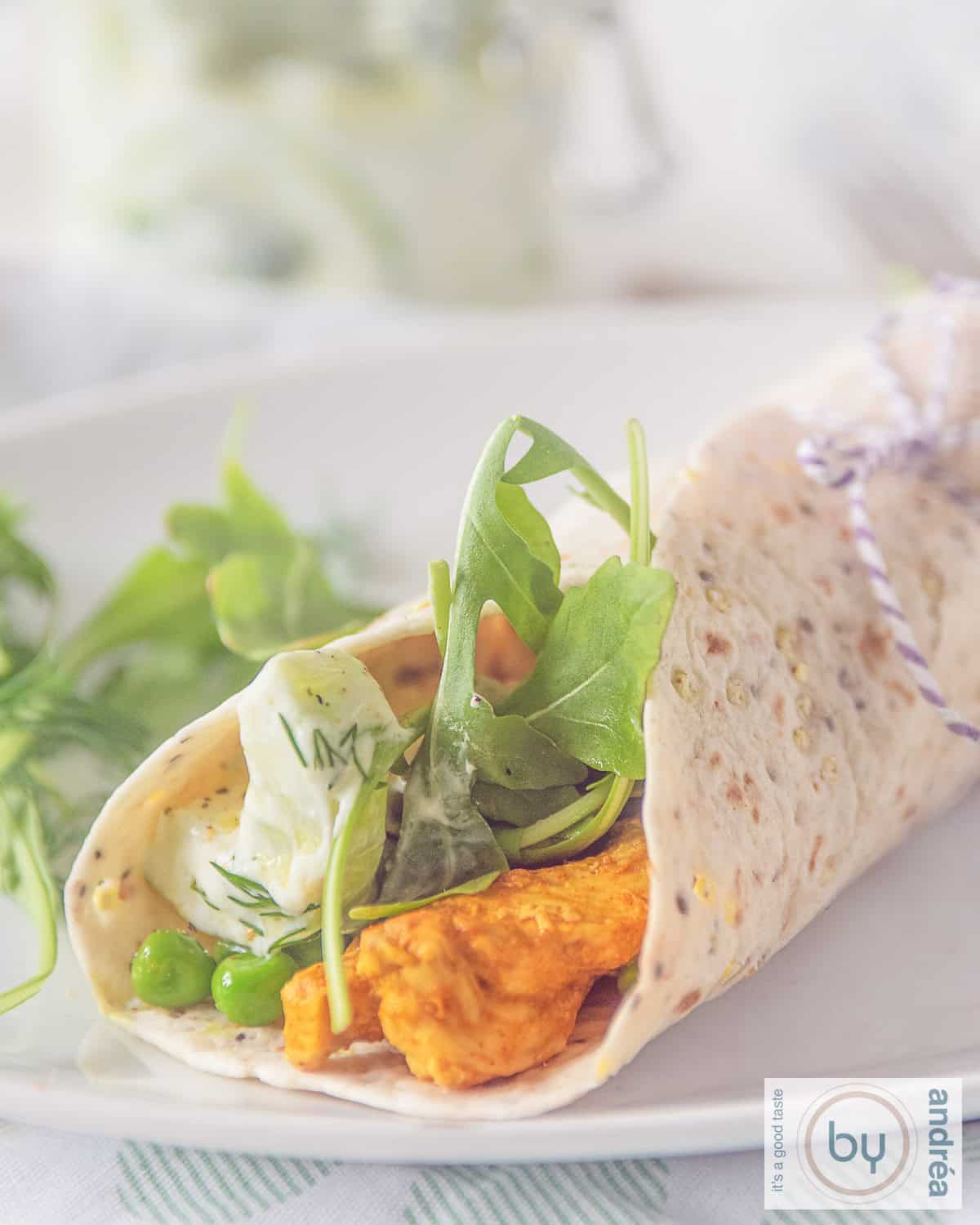 A white plate with a wrap filled with Tandoori chicken, vegetables and tzatziki