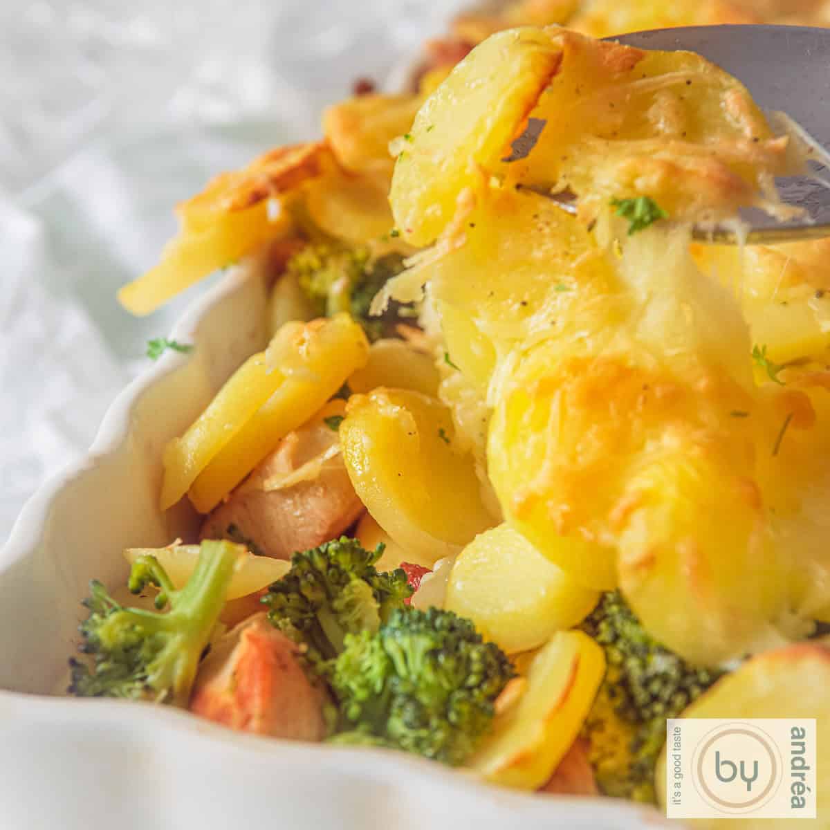 A square photo with part of a white baking dish. A top layer of gratinated potatoes. Below that you see broccoli and chicken.