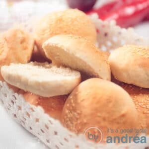 A square shot of a white bowl filled with hamburger buns. One of the sandwiches has been cut open