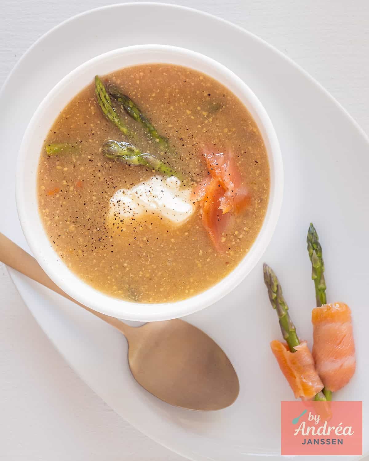 A photo from above of a white platter with a white bowl on top containing mustard soup with asparagus, smoked salmon, and crème fraîche. Two asparagus spears rolled in salmon lie alongside.