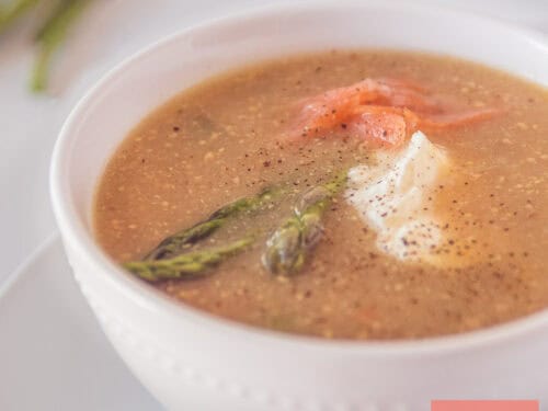 A square photo with a white bowl of mustard soup with green asparagus and salmon