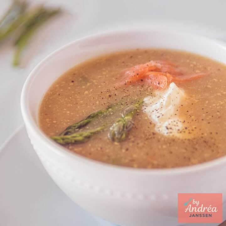 A square photo with a white bowl of mustard soup with green asparagus and salmon