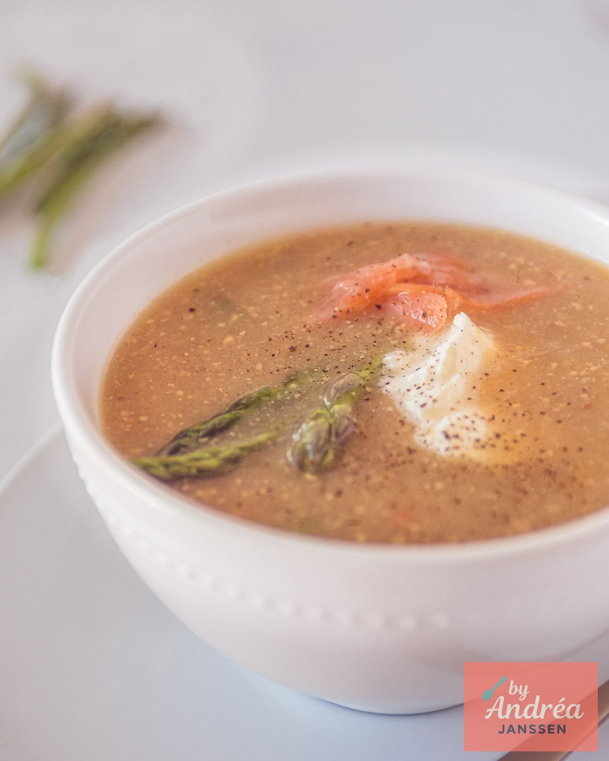 Een kom met zelfgemaakte mosterdsoep, gerookte zalm, groene asperges en crème fraîche op een witte ondergrond.