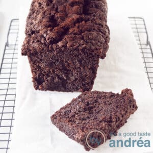 A square photo of a Chocolate zucchini pound cake, one slice cut of laying in front on a baking paper on a rack.