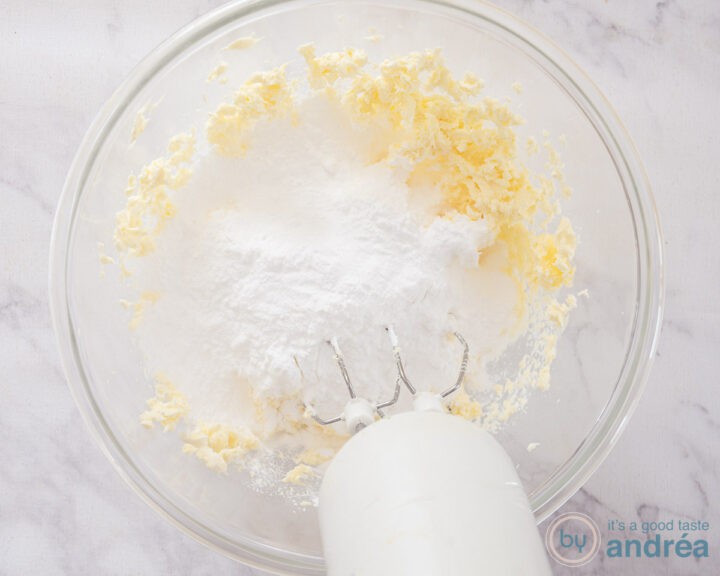A glass bowl with powdered sugar, butter, cream cheese, vanilla and salt and a mixer in it.