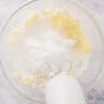 A glass bowl with powdered sugar, butter, cream cheese, vanilla and salt and a mixer in it.