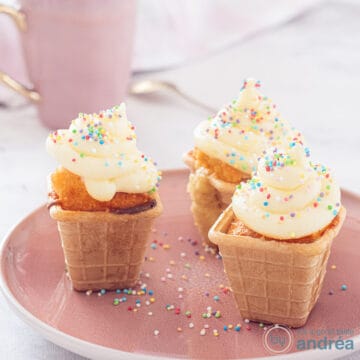 A square photo of 3 ice cream cups of cupcake with frosting on a pink plate.