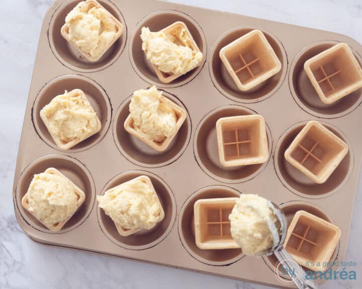 A muffin tin with ice cube trays. Half filled with batter. An ice cream scoop fills an ice cream cup.
