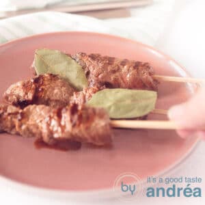 A square photo with a pink plate on which three Portuguese grilled skewers. A hand takes a skewer