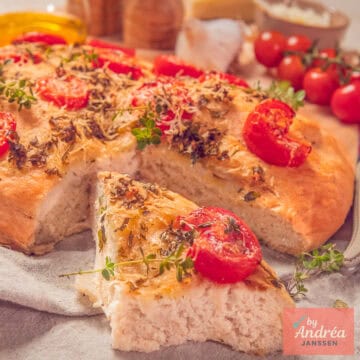 A square photo showing part of a focaccia with a wedge cut out (revealing the airy interior). With a topping of Italian herbs, tomato, garlic, and Parmesan cheese.