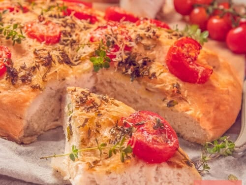 A square photo showing part of a focaccia with a wedge cut out (revealing the airy interior). With a topping of Italian herbs, tomato, garlic, and Parmesan cheese.