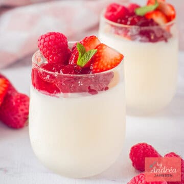 A square photo with two small glasses placed one behind the other with yogurt ice cream, strawberry jam, and red fruit.