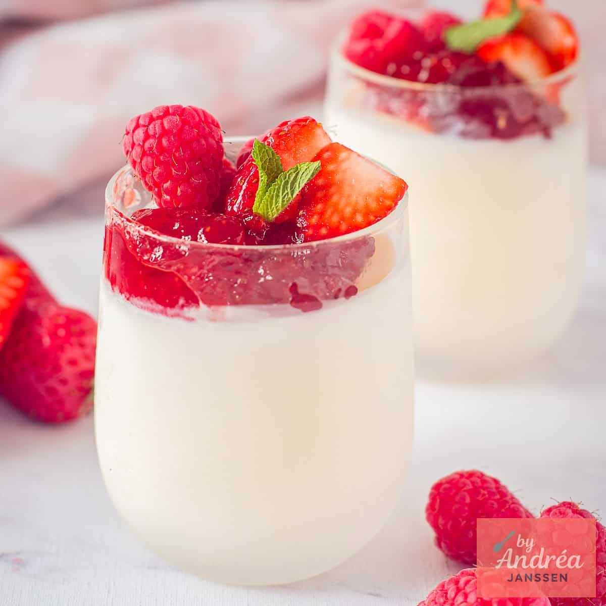 A square photo with two small glasses placed one behind the other with yogurt ice cream, strawberry jam, and red fruit.
