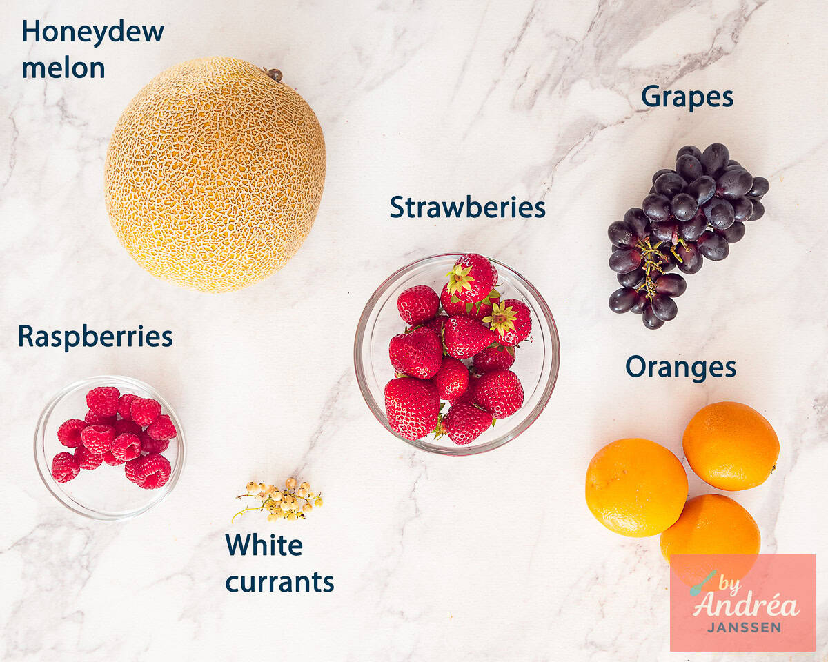 Ingredients Fruit Salad in Orange Cups for Kids on a white marble background with text description.