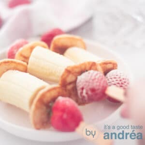 A square photo with three sticks filled with banana, pancakes and strawberries. A hand takes one off the board.